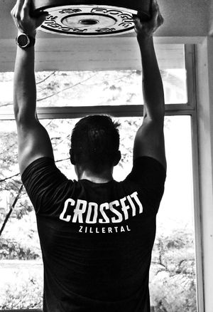 Aufnahme Rückenansicht Athlet mit Crossfit T-Shirt, der mit gestreckten Armen die Platte über dem Kopf hält